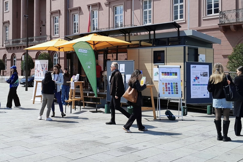 Menschen bei einem Infostand mit Plakaten und Sonnenschirmen auf einem Platz.