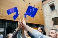 Hands raised together holding multiple European Union flags in an outdoor setting.