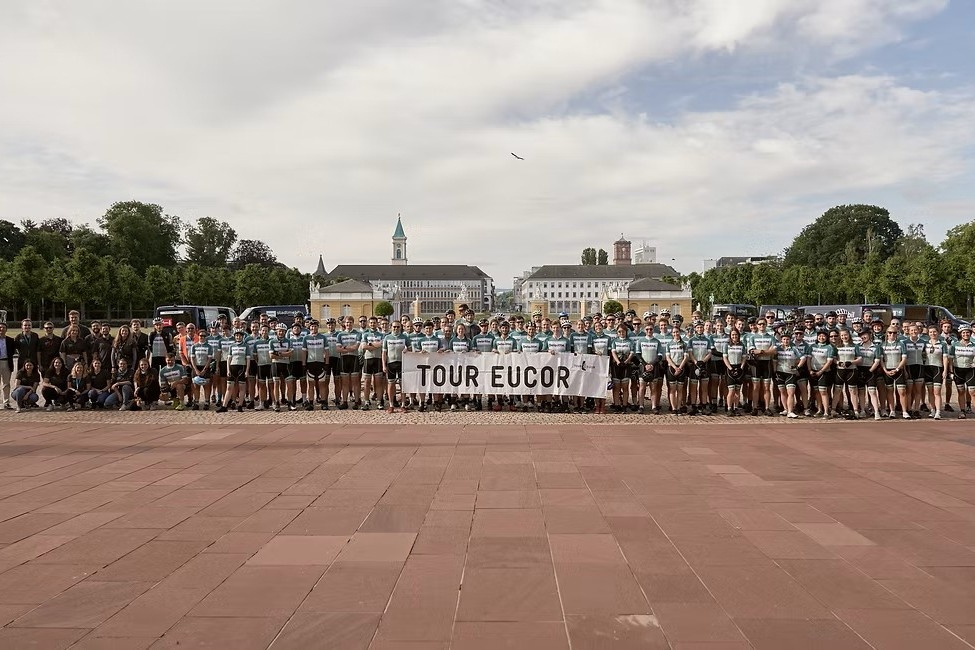 Gruppenfoto von Radfahrern mit Banner Tour EUCOR vor Residenzplatz, Hintergrund Parkanlagen und Gebäude.