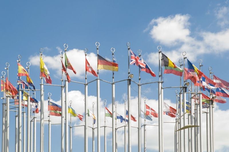 Viele Länderflaggen wehen an hohen Stangen im Freien gegen blauen Himmel.